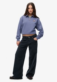 Sudadera corta de color púrpura claro con cremallera y logo en el pecho, combinada con pantalones de pana anchos de color oscuro. Texturas suaves y ajuste relajado.