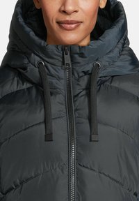 Een vrouw in een zwart gewatteerd pufferjack met een hoge kraag, capuchon en ritssluiting aan de voorkant, waarbij alleen haar ondergezicht en nek zichtbaar zijn.