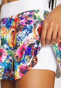 Primer plano de una mano levantando la colorida capa exterior con un patrón abstracto de una falda deportiva multicolor de Nike, revelando unos pantalones cortos blancos interiores.