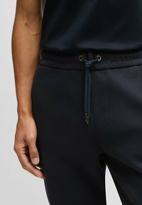 Mörkblå träningsbyxor med dragsko i midjan, som har en strukturerad tyg och metallöglor, visade med en hand som vilar på sidan.