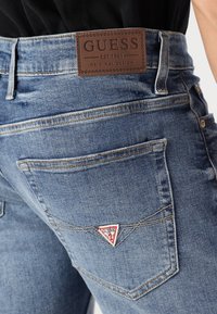 Traperice svijetloplave boje s kožnom oznakom označenom "GUESS" na struku. Karakteristike uključuju stražnje džepove s detaljima u obliku trokuta.