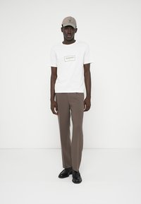 Biela tričko s čiernym logom, hnedé elegantné nohavice, šedá čiapka a čierne topánky. Minimalistický dizajn s hladkou textúrou a čistými líniami.