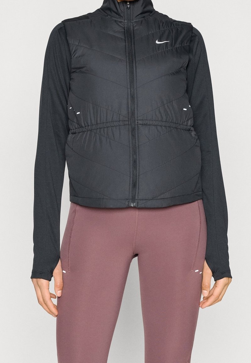 Personne portant une veste matelassée noire Nike zippée par-dessus un t-shirt noir à manches longues et des leggings moulants mauves sur un fond neutre.