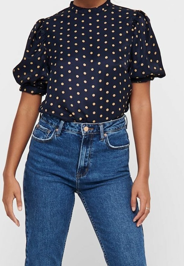 Blouse bleu marine à pois beige avec manches courtes bouffantes assortie à un jean en denim bleu taille haute sur une personne debout.