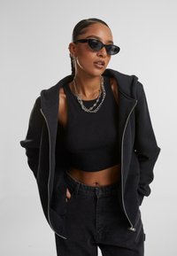 Felpa nera con zip realizzata in tessuto morbido, indossata sopra un crop top nero a coste e abbinata a jeans scuri. Presenta una vestibilità rilassata e accessori in metallo argentato.