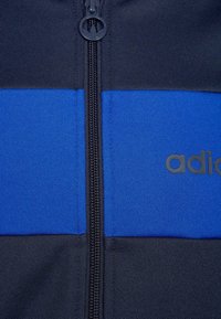 Zip-jacka med ett färgblockdesign i blått och svart. Texturerat tyg med en central dragkedja, logo tryckt i svart på blått.