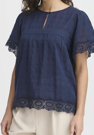 Nainen, joka pukeutuu tummansiniseen paitapuseroon, jossa on pitsireunukset hihoissa ja helmassa, pieni etuleikkaus napilla, yhdistettynä beigeihin housuihin.