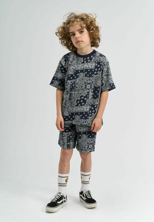 Tumesinise puuvillase komplekti, mis sisaldab lühikeste varrukatega särki ja lühikesi pükse, kaunistatud valge paisley- ja lillemustriga. Mustad spordijalatsid ja triibulised sokid.