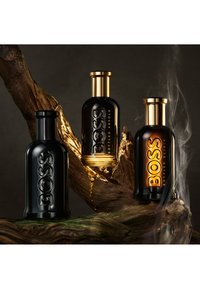 Trois flacons de parfum Boss noirs et dorés disposés sur du bois texturé, l'un émettant de la fumée montante sur un fond sombre.