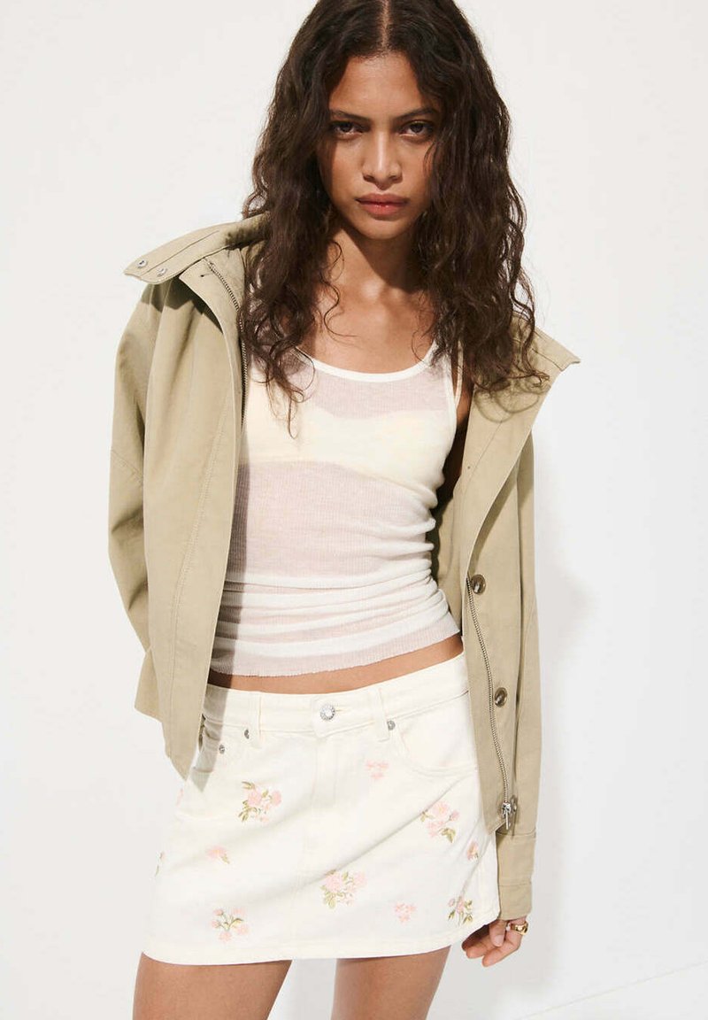Femme aux longs cheveux bouclés portant une veste beige par-dessus un débardeur blanc et une mini-jupe blanche à fleurs, posant devant un fond blanc.