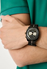 Orologio cronografo nero con cinturino in tessuto testurizzato e accenti dorati, dotato di un quadrante nero con sottoquadranti bianchi e una scala tachimetrica.