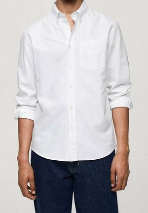 Personne portant une chemise blanche boutonnée avec les manches retroussées et un jean bleu foncé, debout devant un fond clair uni.