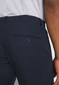 Marineblauwe broek van gestructureerde stof, met een achterzak met een knoop en een subtiele merkaccent. Elastische tailleband zichtbaar.