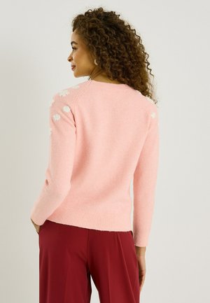 Mujer con cabello rizado que lleva un suéter rosa con detalles de flores blancas en los hombros y pantalones rojos, de perfil contra un fondo liso.