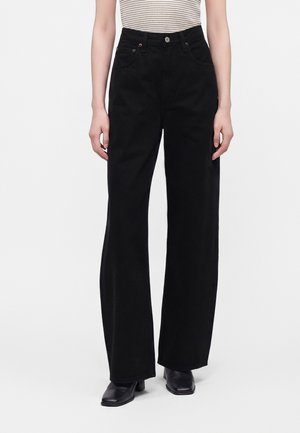 Personne portant un pantalon large noir taille haute, un haut rayé sans manches et des bottines noires à talons, debout devant un fond uni.