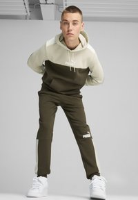 Sudadera de color oliva y crema con un bolsillo tipo canguro, combinada con pantalones oliva que tienen paneles laterales y logotipos en blanco. Zapatillas blancas completan el look.