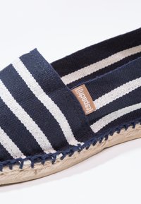 Marineblau und weiße gestreifte Leinensandale mit einer geflochtenen Jutesohle, ausgestattet mit einem braunen Leder-Logoschild an der Seite.