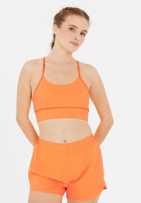 Orange sportbh med tunna axelband och en åtsittande design, parat med orange shorts med elastisk midja som har en lös passform och en mjuk textur.