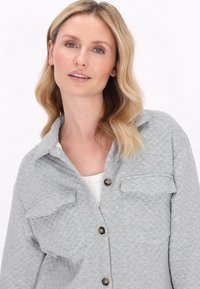 Giacca camicia trapuntata grigia con colletto, due tasche sul petto e bottoni marroni; il tessuto strutturato aggiunge interesse visivo.