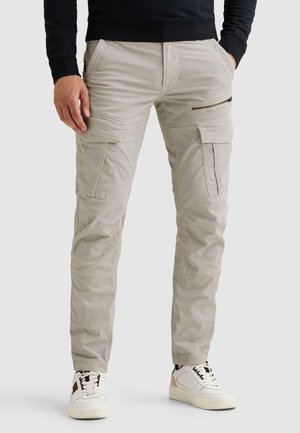 Man die lichtbeige cargobroek met meerdere zakken en witte sneakers draagt, handen half in de zakken tegen een effen achtergrond.
