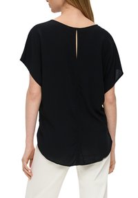 Blusa negra de manga corta con corte holgado, que presenta un detalle de abertura en la parte posterior y una tela suave y ligera, combinada con pantalones de color claro.