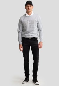 Vīrietis, kas valkā melnu cepuri, pelēku sporta krekla "BIG STAR" virs baltas krekla, melnas džinsas un melnus sporta apavus, stāv pret vienkāršu fonu.