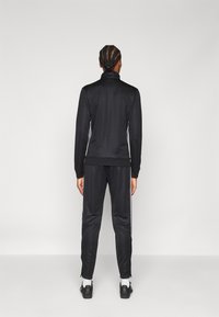 Survêtement de sport noir avec un col haut et des rayures sur les côtés. Texture lisse, design ajusté, doté de poignets élastiques et de chevilles zippées.