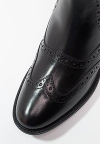 Zapato brogue de cuero negro con punta afilada, perforaciones decorativas y un acabado brillante con suela cosida.