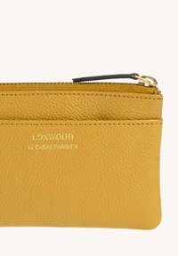 Pochette en cuir texturé jaune avec fermeture éclair dorée et tirant noir, embossée avec "LOXWOOD LE CASAS PARISIEN" en lettres dorées.