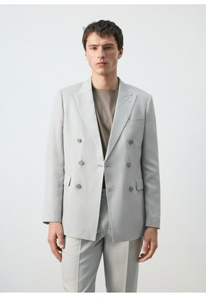 Junger Mann, der einen hellgrauen, doppelt geknöpften Anzug mit einem gedämpften Taupe-Hemd trägt, vor einem schlichten weißen Hintergrund stehend.