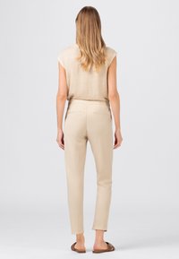 Beige bezrukávový top s volným střihem, kombinovaný se světlými béžovými p�řiléhavými kalhotami a hnědými sandály, pohled zezadu na neutrálním pozadí.