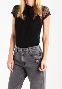 Bodysuit negro con mangas transparentes de patrón estrellado y cuello con lazo, combinado con jeans gris de tiro alto que presentan un detalle de bordado en forma de estrella.