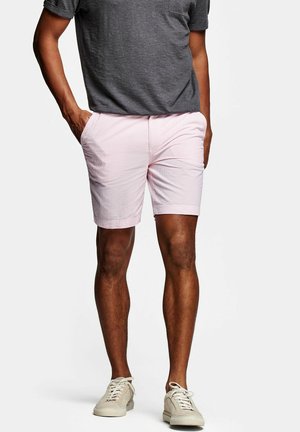 Homme portant un short rose clair, un t-shirt gris foncé et des baskets beiges, se tenant les mains dans les poches devant un fond blanc uni.