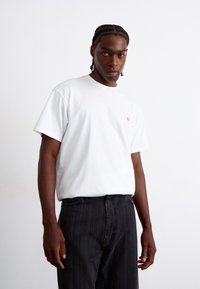 Weißes T-Shirt mit kurzen Ärmeln und einem kleinen gelben Logo auf der Brust, kombiniert mit schwarzen Nadelstreifen-Hosen. Glattes Baumwollgewebe, lässige Passform.