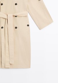 Beige Trenchcoat mit Taillengürtel, strukturiertem Stoff, doppelt geknöpftem Design, schwarzen Knöpfen und zwei Seiten Taschen.