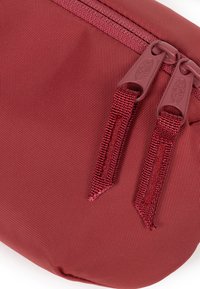 Röd texturerad påse med en slät yta, med dubbla dragkedjor och tygöglor. Kompakt, rundad form för enkel bärbarhet.