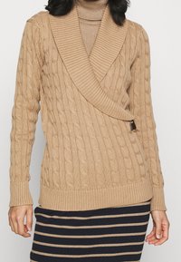 Beżowy sweter z grubym splotem, szerokim kołnierzem typu szalowego, kopertowym krojem, długimi rękawami oraz ozdobą z boku na zapięcie. Wykończenia przy mankietach i na dole swetra są ściągane.