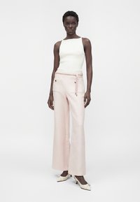 Femme portant un haut blanc sans manches, un pantalon large rose pâle avec poches avant et ceinture à nouer, ainsi que des chaussures blanches à petit talon avec boucles.