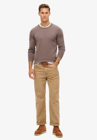 Maglione a maglia color marrone chiaro con texture a coste, abbinato a pantaloni beige e stivali in pelle marrone. Include un dettaglio con colletto bianco.