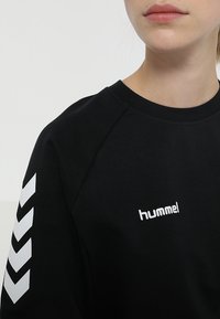 Sweat-shirt noir avec le logo "hummel" blanc sur la poitrine et motif chevron blanc sur la manche. Confectionné en tissu doux avec un col rond.