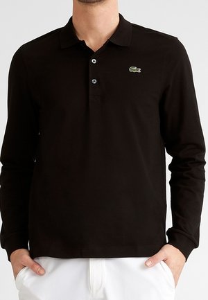 Homme portant un polo noir à manches longues avec deux boutons et un logo crocodile vert sur la poitrine gauche, mains dans les poches d’un pantalon blanc.