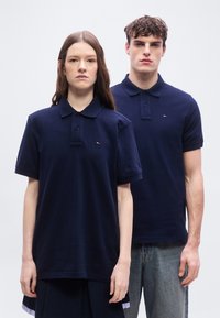 Γυναίκα και άνδρας που στέκονται δίπλα δίπλα φορώντας μπλε μπλουζάκια πόλο με μικρό κόκκινο, λευκό και μπλε λογότυπο στο στήθος, μπροστά σε απλό φόντο.