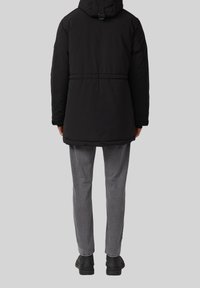 Schwarze isolierte Jacke mit Kapuze, elastischem Bund und gerafften Bündchen, kombiniert mit grauen strukturierten Hosen und schwarzen Schuhen.