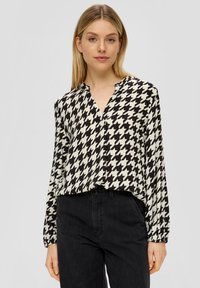 Vrouw in een zwart-wit houndstooth patroon lange mouw blouse, gecombineerd met hoogsluitende zwarte broek, staande tegen een effen achtergrond.