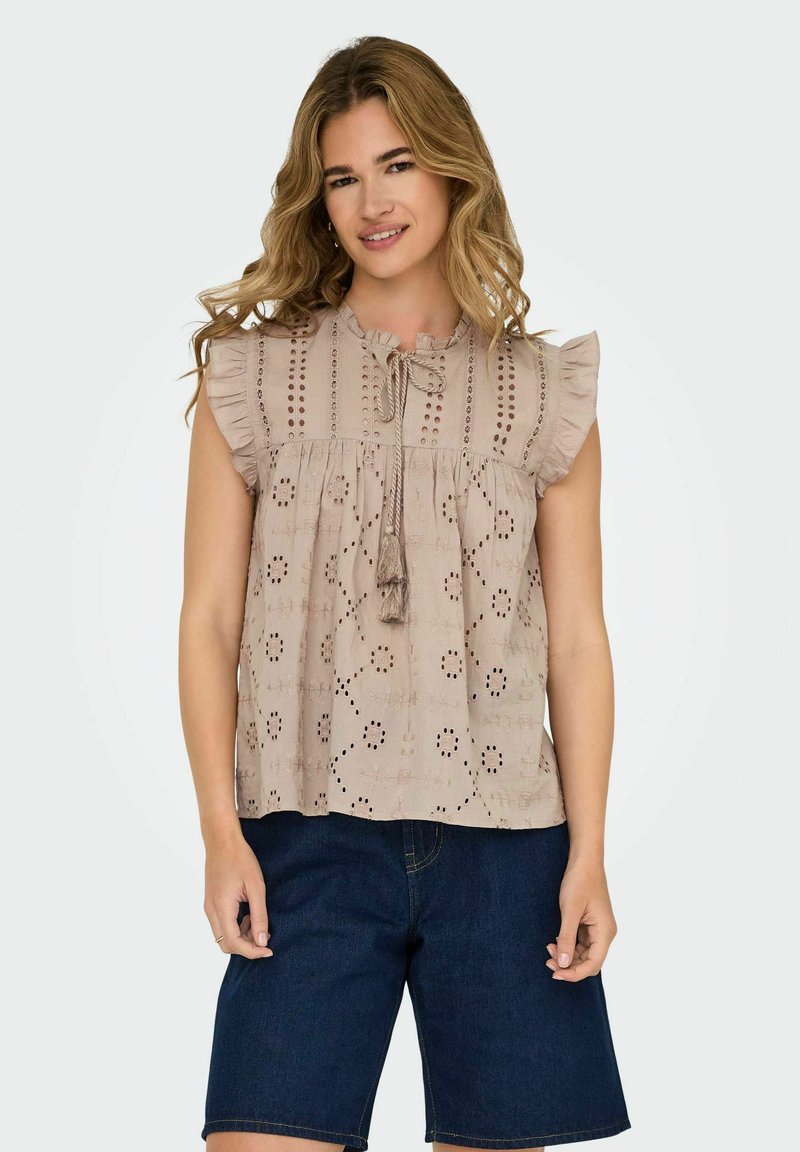 Beige Baumwolloberteil mit Rüschensäumen, das mit Häkelmustern versehen ist und einen gebundenen Rundhalsausschnitt mit Quasten hat. Kombiniert mit dunklen Jeanskurzshorts.