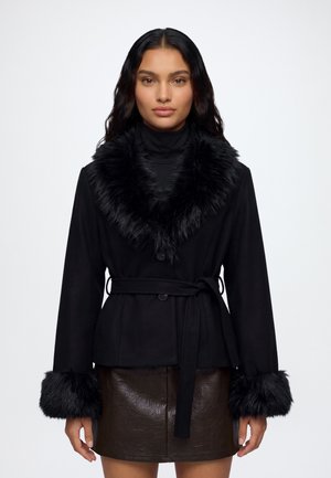 Femme portant un manteau noir ceinturé avec une garniture en fausse fourrure au col et aux poignets, associé à une jupe en cuir marron foncé, sur un fond uni.