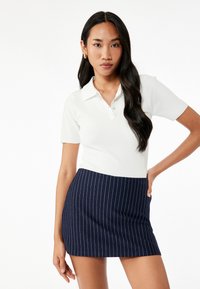 Valkoinen ribattu polo-paita lyhyillä hihoilla ja kolmella napilla, yhdistettynä laivastonsiniseen pinstripe-minihameeseen. Liukas rakenne ja istuva muotoilu.