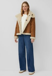 Brązowa zamszowa kurtka z kołnierzem i mankietami z sztucznego futra, ozdobiona dużymi czarnymi guzikami, w zestawieniu z szerokimi niebieskimi dżinsami i kremowym obuwiem.