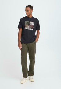 Svart t-shirt med flerfärgad "SHELBY" grafik, kombinerad med olivgröna byxor och vita sneakers. Slät, avslappnad tygkvalitet och avslappnad passform.