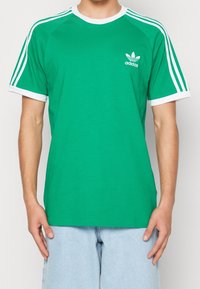 Grön bomulls t-shirt med vit kant och tre vita ränder på varje axel; liten vit Adidas-logotyp på övre vänstra bröstet.